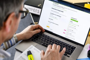 Gros plan sur une personne penchée devant son ordinateur portable (MacBook Pro) : l’écran affiche le formulaire « Déclaration d’impôt 2023 » sur le site de l’État de Vaud. Ses mains tapent au clavier. À côté de l’ordinateur, on aperçoit un classeur noir étiqueté « IMPÔTS », des documents imprimés à en-tête « État de Vaud » et un surligneur jaune.