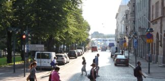 Dank Tempo 30: Helsinki verzeichnet ein Jahr ohne Verkehrstote Mehrere Fussgänger:innen queren an einem sonnigen Nachmittag eine breite, mit Kopfsteinpflaster ausgelegte Strasse in Helsinki, Finnland. Im Vordergrund laufen fünf Personen in unterschiedlichen Richtungen – teils allein, teils in kleinen Abständen zueinander. Links schliesst sich ein schattiger, von hohen Bäumen gesäumter Parkbereich an, teils verdeckt von geparkten Autos und einem Verkehrsschild mit den Aufschriften „Kluuvi“ und „Kasarmitori“. Rechts reihen sich historische mehrstöckige Gebäude mit steinernen Fassaden aneinander, davor weitere parkende Fahrzeuge und Hinweisschilder. Im Hintergrund öffnen sich die breiten Strassenzüge weiter ins Helle, wo man am Horizont weitere Personen, Autos und städtisches Leben erahnt.