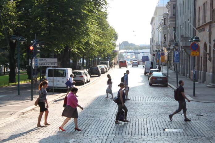 Mehrere Fussgänger:innen queren an einem sonnigen Nachmittag eine breite, mit Kopfsteinpflaster ausgelegte Strasse in Helsinki, Finnland. Im Vordergrund laufen fünf Personen in unterschiedlichen Richtungen – teils allein, teils in kleinen Abständen zueinander. Links schliesst sich ein schattiger, von hohen Bäumen gesäumter Parkbereich an, teils verdeckt von geparkten Autos und einem Verkehrsschild mit den Aufschriften „Kluuvi“ und „Kasarmitori“. Rechts reihen sich historische mehrstöckige Gebäude mit steinernen Fassaden aneinander, davor weitere parkende Fahrzeuge und Hinweisschilder. Im Hintergrund öffnen sich die breiten Strassenzüge weiter ins Helle, wo man am Horizont weitere Personen, Autos und städtisches Leben erahnt.