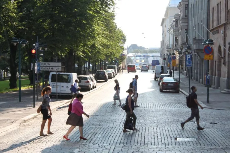 Mehrere Fussgänger:innen queren an einem sonnigen Nachmittag eine breite, mit Kopfsteinpflaster ausgelegte Strasse in Helsinki, Finnland. Im Vordergrund laufen fünf Personen in unterschiedlichen Richtungen – teils allein, teils in kleinen Abständen zueinander. Links schliesst sich ein schattiger, von hohen Bäumen gesäumter Parkbereich an, teils verdeckt von geparkten Autos und einem Verkehrsschild mit den Aufschriften „Kluuvi“ und „Kasarmitori“. Rechts reihen sich historische mehrstöckige Gebäude mit steinernen Fassaden aneinander, davor weitere parkende Fahrzeuge und Hinweisschilder. Im Hintergrund öffnen sich die breiten Strassenzüge weiter ins Helle, wo man am Horizont weitere Personen, Autos und städtisches Leben erahnt.
