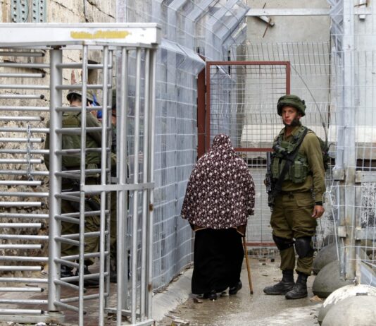 Druck auf Israel: Wieso die Zweistaatenlösung wichtig ist Eine Frau mit langem schwarzem Gewand und braun-weißem Blumenschal über dem Kopf stützt sich auf einen Gehstock und geht durch einen engen Korridor aus hohen Metallzäunen. Links im Bild sieht man ein drehkreuzartiges Tor, dahinter einen in olivgrüner Uniform und Helm gekleideten Soldaten. Rechts steht ein weiterer Soldat in Kampfmontur mit Helm und Gewehr, der in Richtung der Kamera blickt. Der Boden ist feucht und mehrere große Betonkugeln sichern die Barrieren an den Seiten.