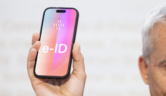 Ein Mann hält ein Smartphone hoch, auf dessen Bildschirm gross ‹e-ID› steht. Die Darstellung symbolisiert die geplante elektronische Identität der Schweiz