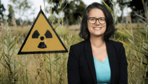 Une femme portant des lunettes, un blazer noir et un T-shirt bleu clair sourit devant un panneau triangulaire jaune arborant le symbole radioactif, le tout planté dans des hautes herbes en milieu naturel.