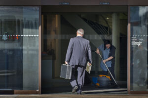 Photo couleur d’une entrée vitrée d’une agence UBS. Un homme d’affaires en costume, vu de dos, franchit le seuil en tenant une mallette noire dans une main et un journal dans l’autre. À droite, une employée de nettoyage en uniforme passe l’aspirateur sur le paillasson. Le logo UBS apparaît en rouge sur la porte en verre.