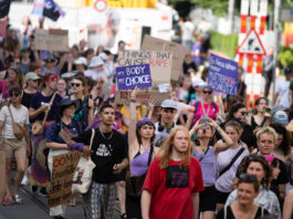 Good News im September – von lokal bis hin zur Erdatmosphäre Vue d’une manifestation en plein air où un groupe divers de personnes marche dans la rue. Au centre, une jeune femme en haut violet lève une pancarte « My body, my choice ». Plusieurs manifestant·e·s autour portent des vêtements et accessoires violets et tiennent d’autres slogans (dont, en arrière-plan, « Things that cause rape : »). L’ambiance est estivale, la foule avance unie pour défendre les droits à l’avortement et l’autodétermination.