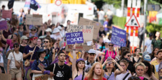 Good News im September – von lokal bis hin zur Erdatmosphäre Vue d’une manifestation en plein air où un groupe divers de personnes marche dans la rue. Au centre, une jeune femme en haut violet lève une pancarte « My body, my choice ». Plusieurs manifestant·e·s autour portent des vêtements et accessoires violets et tiennent d’autres slogans (dont, en arrière-plan, « Things that cause rape : »). L’ambiance est estivale, la foule avance unie pour défendre les droits à l’avortement et l’autodétermination.