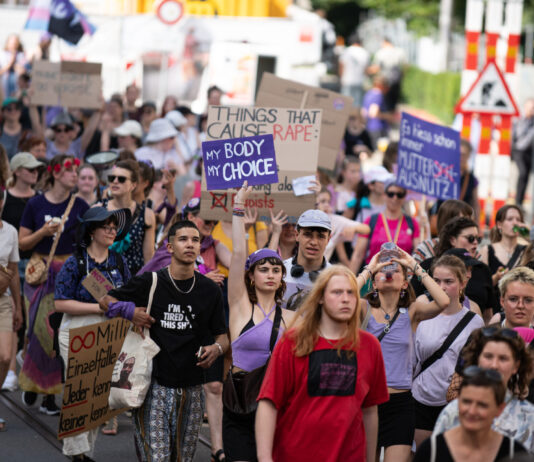 Good News im September – von lokal bis hin zur Erdatmosphäre Vue d’une manifestation en plein air où un groupe divers de personnes marche dans la rue. Au centre, une jeune femme en haut violet lève une pancarte « My body, my choice ». Plusieurs manifestant·e·s autour portent des vêtements et accessoires violets et tiennent d’autres slogans (dont, en arrière-plan, « Things that cause rape : »). L’ambiance est estivale, la foule avance unie pour défendre les droits à l’avortement et l’autodétermination.