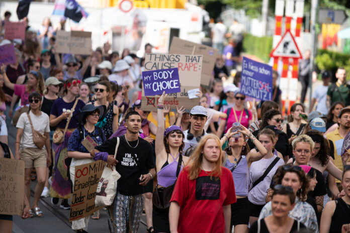Vue d’une manifestation en plein air où un groupe divers de personnes marche dans la rue. Au centre, une jeune femme en haut violet lève une pancarte « My body, my choice ». Plusieurs manifestant·e·s autour portent des vêtements et accessoires violets et tiennent d’autres slogans (dont, en arrière-plan, « Things that cause rape : »). L’ambiance est estivale, la foule avance unie pour défendre les droits à l’avortement et l’autodétermination.