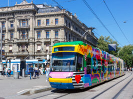 365-Franken-Initiative: Ein Schlüssel zur Mobilitätswende Bunte ZVV-Straßenbahn (Linie 11 „Rehalp“) vor einem historischen, mit „Credit Suisse“ beschrifteten Gebäude, im Hintergrund wartende Passanten und Trambahnen unter klarem blauem Himmel.