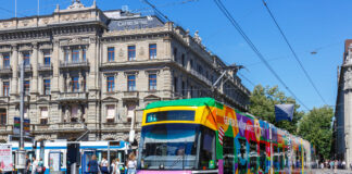365-Franken-Initiative: Ein Schlüssel zur Mobilitätswende Bunte ZVV-Straßenbahn (Linie 11 „Rehalp“) vor einem historischen, mit „Credit Suisse“ beschrifteten Gebäude, im Hintergrund wartende Passanten und Trambahnen unter klarem blauem Himmel.