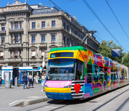 365-Franken-Initiative: Ein Schlüssel zur Mobilitätswende Bunte ZVV-Straßenbahn (Linie 11 „Rehalp“) vor einem historischen, mit „Credit Suisse“ beschrifteten Gebäude, im Hintergrund wartende Passanten und Trambahnen unter klarem blauem Himmel.