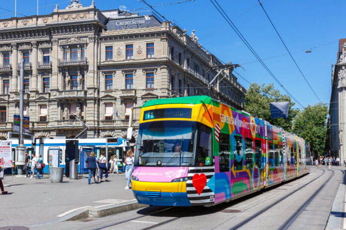 Bunte ZVV-Straßenbahn (Linie 11 „Rehalp“) vor einem historischen, mit „Credit Suisse“ beschrifteten Gebäude, im Hintergrund wartende Passanten und Trambahnen unter klarem blauem Himmel.