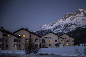 Vue de nuit tombante d’un petit ensemble de maisons beiges aux toits foncés à Sils, aux Grisons, dont plusieurs fenêtres sont baignées d’une lumière chaude. Elles se dressent au-dessus d’un champ enneigé, devant le flanc d’une haute montagne également couverte de neige, sous un ciel bleu sombre de crépuscule.