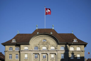 Façade de la Banque nationale suisse à Berne, bâtiment classique à toiture en tuiles brunes, drapeau suisse flottant sur un mât central, ciel bleu en arrière-plan.