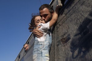 Sous un ciel bleu, un homme penché par la fenêtre d’un autocar sombre serre une petite fille dans ses bras et l’embrasse sur la joue. La fillette, vêtue d’une salopette en jean et d’un t-shirt blanc, agrippe le rebord du véhicule, le regard mélancolique, tandis qu’un autre passager observe la scène en arrière-plan.
