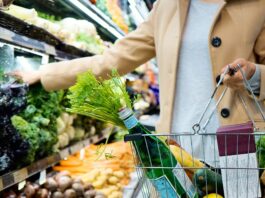Nestlé, Coca-Cola und Co.: Ein Blick auf drei Riesen im globalen Lebensmittelmarkt Eine Person in einem beigen Mantel steht im Supermarkt vor einer Gemüsetheke. Sie hält einen drahtfertigen Einkaufskorb mit einer grünen Mineralwasserflasche, frischem Karottengrün, einer Zitrone, einer Zucchini, einem Baguette und zwei verpackten Lebensmitteln, während sie mit der anderen Hand nach Kohl und Brokkoli greift.
