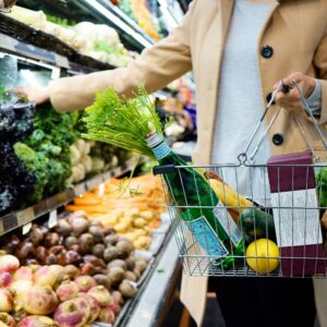 Dans une épicerie, une personne vêtue d’un manteau beige tient un panier métallique rempli d’une bouteille d’eau gazeuse, d’un bouquet de fanes de carottes, d’un citron, d’une courgette et de quelques produits emballés. Elle se tient devant les étals garnis de choux, de brocolis, de betteraves, de navets et de carottes.