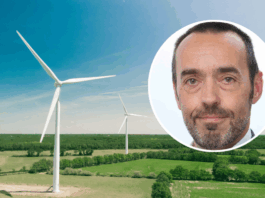Bruno Tobback: «Der ‹Green Deal› ist auch eine Unabhängigkeitserklärung» Luftaufnahme einer weitläufigen, grünen Landschaft mit zwei großen weissen Windrädern unter einem klaren blauen Himmel. Oben rechts ist ein kreisförmiges Bild überlagert, das das Gesicht von Bruno Tobback, einem Mann mittleren Alters zeigt, der in die Kamera blickt, kurze Haare hat und einen leichten grauen Bart trägt.