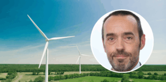 Bruno Tobback: «Der ‹Green Deal› ist auch eine Unabhängigkeitserklärung» Luftaufnahme einer weitläufigen, grünen Landschaft mit zwei großen weissen Windrädern unter einem klaren blauen Himmel. Oben rechts ist ein kreisförmiges Bild überlagert, das das Gesicht von Bruno Tobback, einem Mann mittleren Alters zeigt, der in die Kamera blickt, kurze Haare hat und einen leichten grauen Bart trägt.