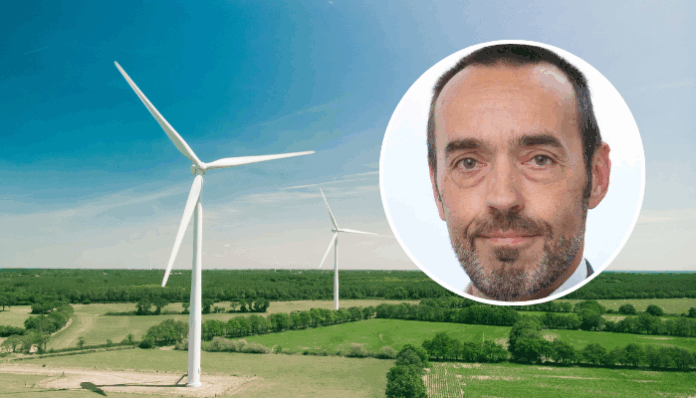 Tobback Luftaufnahme einer weitläufigen, grünen Landschaft mit zwei großen weissen Windrädern unter einem klaren blauen Himmel. Oben rechts ist ein kreisförmiges Bild überlagert, das das Gesicht von Bruno Tobback, einem Mann mittleren Alters zeigt, der in die Kamera blickt, kurze Haare hat und einen leichten grauen Bart trägt.