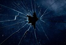 Femizide: Die unbequeme Wahrheit Nahaufnahme einer dunklen zerbrochenen Glasscheibe, bei der sternförmige Risse von einem zentralen Loch ausgehen.