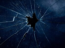 Femizide: Die unbequeme Wahrheit Nahaufnahme einer dunklen zerbrochenen Glasscheibe, bei der sternförmige Risse von einem zentralen Loch ausgehen.