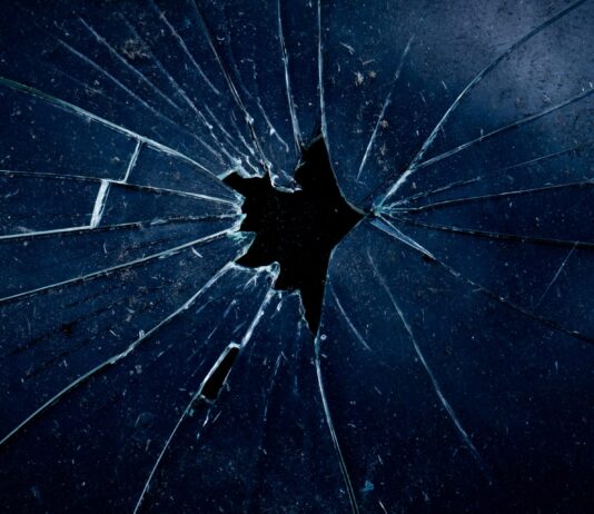 Femizide: Die unbequeme Wahrheit Nahaufnahme einer dunklen zerbrochenen Glasscheibe, bei der sternförmige Risse von einem zentralen Loch ausgehen.