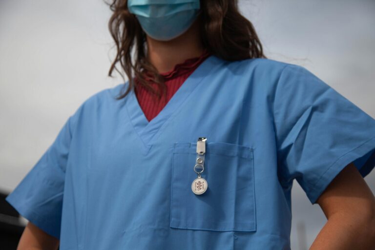 Nahaufnahme des Oberkörpers einer Pflegefachperson in blauem Kittel und chirurgischer Maske. An der Brusttasche ist ein „Medical Alert“-Button befestigt, darunter schimmert ein bordeauxrotes Oberteil hervor.
