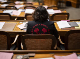 Eine halbe Million Unterschriften: Wenn Frauen sich mobilisieren Rückansicht von Tamara Funiciello, die in einem Halbkreis mit Holzmöbeln sitzt, vor ihr Reihen von Schreibtischen, die mit Akten, Blättern und einem Laptop übersät sind. Sie trägt eine schwarze Jacke mit einer roten Aufschrift „Ni Una Menos” (Nicht eine weniger), die zwei stilisierte Rosen umgibt.
