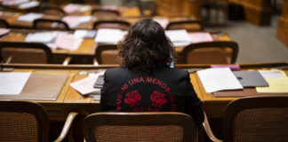 Eine halbe Million Unterschriften: Wenn Frauen sich mobilisieren Rückansicht von Tamara Funiciello, die in einem Halbkreis mit Holzmöbeln sitzt, vor ihr Reihen von Schreibtischen, die mit Akten, Blättern und einem Laptop übersät sind. Sie trägt eine schwarze Jacke mit einer roten Aufschrift „Ni Una Menos” (Nicht eine weniger), die zwei stilisierte Rosen umgibt.