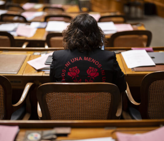 Eine halbe Million Unterschriften: Wenn Frauen sich mobilisieren Rückansicht von Tamara Funiciello, die in einem Halbkreis mit Holzmöbeln sitzt, vor ihr Reihen von Schreibtischen, die mit Akten, Blättern und einem Laptop übersät sind. Sie trägt eine schwarze Jacke mit einer roten Aufschrift „Ni Una Menos” (Nicht eine weniger), die zwei stilisierte Rosen umgibt.
