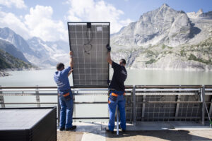 Deux techniciens en tenue de travail bleue soulèvent et positionnent un grand panneau solaire contre une rambarde métallique, avec derrière eux un vaste réservoir d’eau d’un vert pâle encerclé de falaises et de sommets rocheux sous un ciel parsemé de nuages.