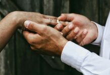 Individualbesteuerung: Ein Steuersystem für mehr Gleichstellung Nahaufnahme zweier Hände: Eine Person in weissem Hemd steckt einer anderen, die bereits einen feinen Diamantring am Ringfinger trägt, einen schlichten silbernen Ring auf. Im unscharfen Hintergrund sind dunkelgraue Holzbretter zu erkennen.