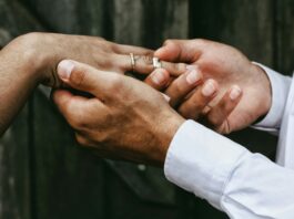 Individualbesteuerung: Ein Steuersystem für mehr Gleichstellung Nahaufnahme zweier Hände: Eine Person in weissem Hemd steckt einer anderen, die bereits einen feinen Diamantring am Ringfinger trägt, einen schlichten silbernen Ring auf. Im unscharfen Hintergrund sind dunkelgraue Holzbretter zu erkennen.