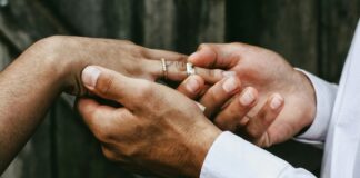 Individualbesteuerung: Ein Steuersystem für mehr Gleichstellung Nahaufnahme zweier Hände: Eine Person in weissem Hemd steckt einer anderen, die bereits einen feinen Diamantring am Ringfinger trägt, einen schlichten silbernen Ring auf. Im unscharfen Hintergrund sind dunkelgraue Holzbretter zu erkennen.