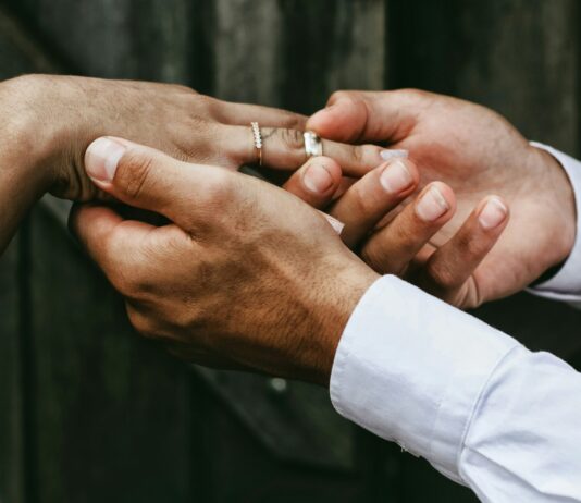 Individualbesteuerung: Ein Steuersystem für mehr Gleichstellung Nahaufnahme zweier Hände: Eine Person in weissem Hemd steckt einer anderen, die bereits einen feinen Diamantring am Ringfinger trägt, einen schlichten silbernen Ring auf. Im unscharfen Hintergrund sind dunkelgraue Holzbretter zu erkennen.