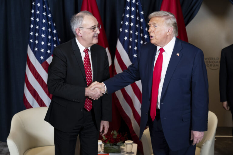 Zwei Männer, Guy Parmelin und Donald Trump, in Anzug und Krawatte geben sich vor mehreren Flaggen (USA und Schweiz) die Hand. Der eine trägt ein dunkelblaues Sakko mit roter Krawatte, der andere einen dunklen Anzug mit gemusterter roter Krawatte. Im Hintergrund stehen zwei weiße Sessel und ein kleiner Tisch mit Blumenarrangement und Wassergläsern.