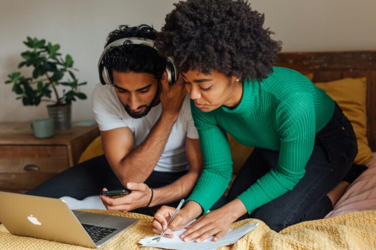 Zwei Personen sitzen auf einem mit einer senfgelben Decke bezogenen Bett. Ein Mann mit schwarzem, lockigem Haar und weißem T-Shirt trägt Kopfhörer und schaut auf sein Smartphone. Daneben lehnt sich eine Frau mit kurzem, krausem Haar und grünem Rippstrickpullover vor, um mit einem Kugelschreiber Notizen auf einem Blatt Papier zu machen. Vor ihnen steht ein aufgeklappter, silberner Laptop, im Hintergrund auf einem hölzernen Nachttisch eine Topfpflanze und eine Kaffeetasse.