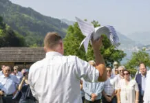 14. Juni: Extremes SVP-Programm könnte umgesetzt werden Versammlung der SVP auf der Rütli-Wiese: Ein Mann in weißem Hemd hält ein weißes Segelflugzeug-Modell in die Höhe, während er vor einer Gruppe von Menschen an einem Bergsee steht. Im Hintergrund sind bewaldete Berge, traditionelle Chalets und ein glitzernder See sichtbar.