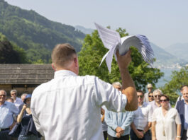 14. Juni: Extremes SVP-Programm könnte umgesetzt werden Versammlung der SVP auf der Rütli-Wiese: Ein Mann in weißem Hemd hält ein weißes Segelflugzeug-Modell in die Höhe, während er vor einer Gruppe von Menschen an einem Bergsee steht. Im Hintergrund sind bewaldete Berge, traditionelle Chalets und ein glitzernder See sichtbar.