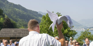 14. Juni: Extremes SVP-Programm könnte umgesetzt werden Versammlung der SVP auf der Rütli-Wiese: Ein Mann in weißem Hemd hält ein weißes Segelflugzeug-Modell in die Höhe, während er vor einer Gruppe von Menschen an einem Bergsee steht. Im Hintergrund sind bewaldete Berge, traditionelle Chalets und ein glitzernder See sichtbar.