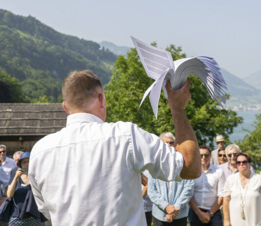 14. Juni: Extremes SVP-Programm könnte umgesetzt werden Versammlung der SVP auf der Rütli-Wiese: Ein Mann in weißem Hemd hält ein weißes Segelflugzeug-Modell in die Höhe, während er vor einer Gruppe von Menschen an einem Bergsee steht. Im Hintergrund sind bewaldete Berge, traditionelle Chalets und ein glitzernder See sichtbar.
