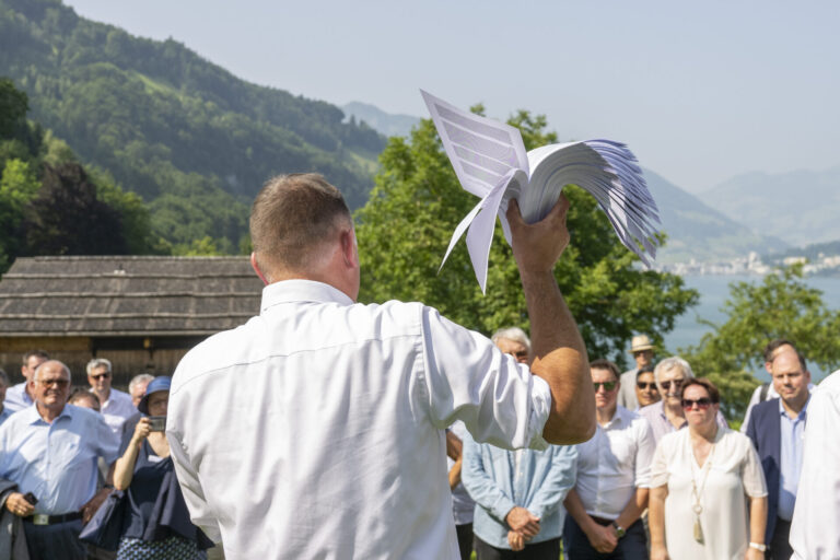Versammlung der SVP auf der Rütli-Wiese: Ein Mann in weißem Hemd hält ein weißes Segelflugzeug-Modell in die Höhe, während er vor einer Gruppe von Menschen an einem Bergsee steht. Im Hintergrund sind bewaldete Berge, traditionelle Chalets und ein glitzernder See sichtbar.