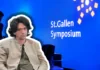 Curtis Yarvin – Vordenker der autoritären Rechten in St. Gallen Farbfotografie einer Präsentationsszene beim St.Gallen Symposium. Im Vordergrund sitzt ein etwa vierzigjähriger Mann mit braunem Haar, Schulterlänge, Brille, beige heller Jacke und graugrüner Krawatte, frontal zur Kamera. Im unscharfen Hintergrund steht ein Mann im blauen Anzug. Der Hintergrund ist leuchtendes Blau mit weißem Text „St.Gallen Symposium" und gelben geometrischen Grafikelementen, typisch für die visuelle Identität dieses internationalen Events.