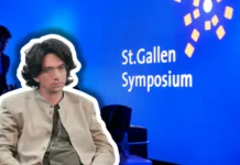 Curtis Yarvin – Vordenker der autoritären Rechten in St. Gallen Farbfotografie einer Präsentationsszene beim St.Gallen Symposium. Im Vordergrund sitzt ein etwa vierzigjähriger Mann mit braunem Haar, Schulterlänge, Brille, beige heller Jacke und graugrüner Krawatte, frontal zur Kamera. Im unscharfen Hintergrund steht ein Mann im blauen Anzug. Der Hintergrund ist leuchtendes Blau mit weißem Text „St.Gallen Symposium" und gelben geometrischen Grafikelementen, typisch für die visuelle Identität dieses internationalen Events.