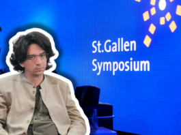 Curtis Yarvin – Vordenker der autoritären Rechten in St. Gallen Farbfotografie einer Präsentationsszene beim St.Gallen Symposium. Im Vordergrund sitzt ein etwa vierzigjähriger Mann mit braunem Haar, Schulterlänge, Brille, beige heller Jacke und graugrüner Krawatte, frontal zur Kamera. Im unscharfen Hintergrund steht ein Mann im blauen Anzug. Der Hintergrund ist leuchtendes Blau mit weißem Text „St.Gallen Symposium" und gelben geometrischen Grafikelementen, typisch für die visuelle Identität dieses internationalen Events.