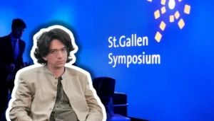 Farbfotografie einer Präsentationsszene beim St.Gallen Symposium. Im Vordergrund sitzt ein etwa vierzigjähriger Mann mit braunem Haar, Schulterlänge, Brille, beige heller Jacke und graugrüner Krawatte, frontal zur Kamera. Im unscharfen Hintergrund steht ein Mann im blauen Anzug. Der Hintergrund ist leuchtendes Blau mit weißem Text „St.Gallen Symposium" und gelben geometrischen Grafikelementen, typisch für die visuelle Identität dieses internationalen Events.