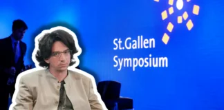 Curtis Yarvin – Vordenker der autoritären Rechten in St. Gallen Farbfotografie einer Präsentationsszene beim St.Gallen Symposium. Im Vordergrund sitzt ein etwa vierzigjähriger Mann mit braunem Haar, Schulterlänge, Brille, beige heller Jacke und graugrüner Krawatte, frontal zur Kamera. Im unscharfen Hintergrund steht ein Mann im blauen Anzug. Der Hintergrund ist leuchtendes Blau mit weißem Text „St.Gallen Symposium" und gelben geometrischen Grafikelementen, typisch für die visuelle Identität dieses internationalen Events.