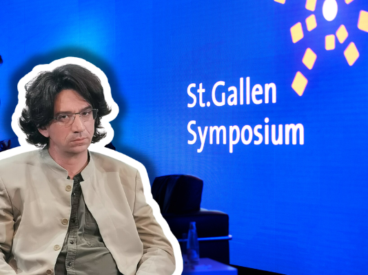 Curtis Yarvin – Vordenker der autoritären Rechten in St. Gallen Farbfotografie einer Präsentationsszene beim St.Gallen Symposium. Im Vordergrund sitzt ein etwa vierzigjähriger Mann mit braunem Haar, Schulterlänge, Brille, beige heller Jacke und graugrüner Krawatte, frontal zur Kamera. Im unscharfen Hintergrund steht ein Mann im blauen Anzug. Der Hintergrund ist leuchtendes Blau mit weißem Text „St.Gallen Symposium" und gelben geometrischen Grafikelementen, typisch für die visuelle Identität dieses internationalen Events.