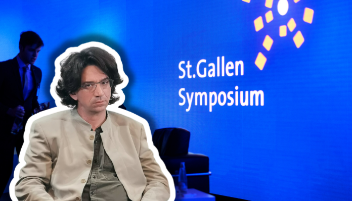 Farbfotografie einer Präsentationsszene beim St.Gallen Symposium. Im Vordergrund sitzt ein etwa vierzigjähriger Mann mit braunem Haar, Schulterlänge, Brille, beige heller Jacke und graugrüner Krawatte, frontal zur Kamera. Im unscharfen Hintergrund steht ein Mann im blauen Anzug. Der Hintergrund ist leuchtendes Blau mit weißem Text „St.Gallen Symposium" und gelben geometrischen Grafikelementen, typisch für die visuelle Identität dieses internationalen Events.